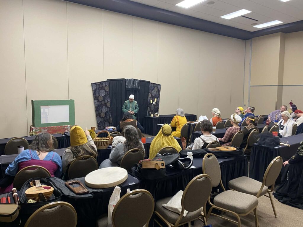 A man in green with a white cap stands in front of a dark backdrop performing before a group of people in historic garb sitting at tables in a conference room.