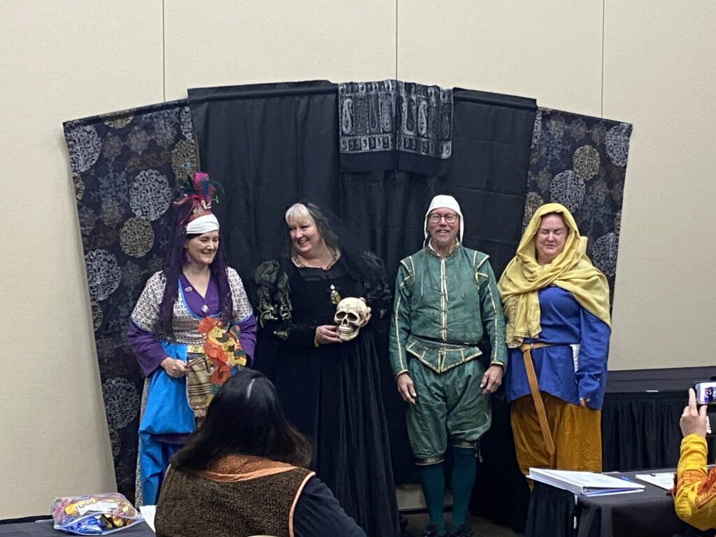 Four people in historic garb stand in front of a dark backdrop. One woman is in colorful Turkish Ottoman garb and holds a paper puppet. The next woman is in a black dress with a black veil and holds a skull. The man next to her is in a green tunic and pants with green stockings and a white cap. The last woman is in a blue tunic with brown pants and a yellow wrap around her head and shoulders.