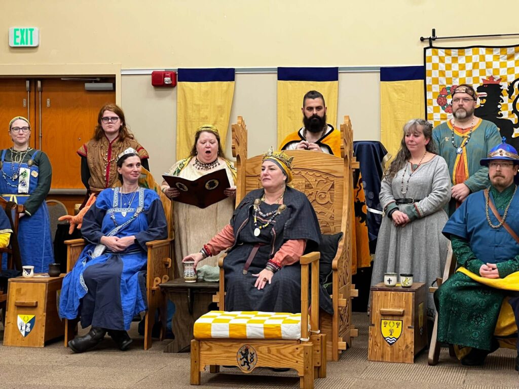 The Queen sits on a wooden throne in a dark blue dress with a rust underlayer. There are two other thrones visible to her sides, one with a Princess in blue and one with a Baron in dark blue and green. Several people stand behind them, including a herald in a natural fabric with her mouth open.