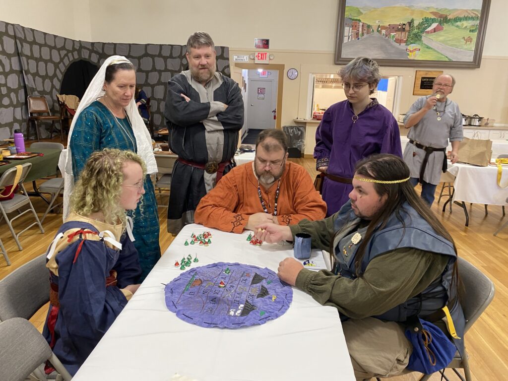Three people are seated around a table playing a game for Hershey kisses. Three more stand around watching them. There is a man in the background eating something and looking their way.